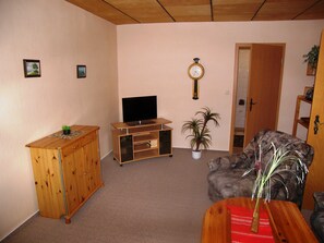 A living room with a wooden cabinet, a television on a wooden stand, a clock on the wall, and a door leading to another room.