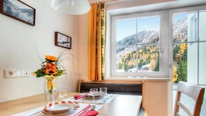 Un coin repas avec une table dressée pour deux personnes, un vase rempli de fleurs et une fenêtre donnant sur une vue sur les montagnes.