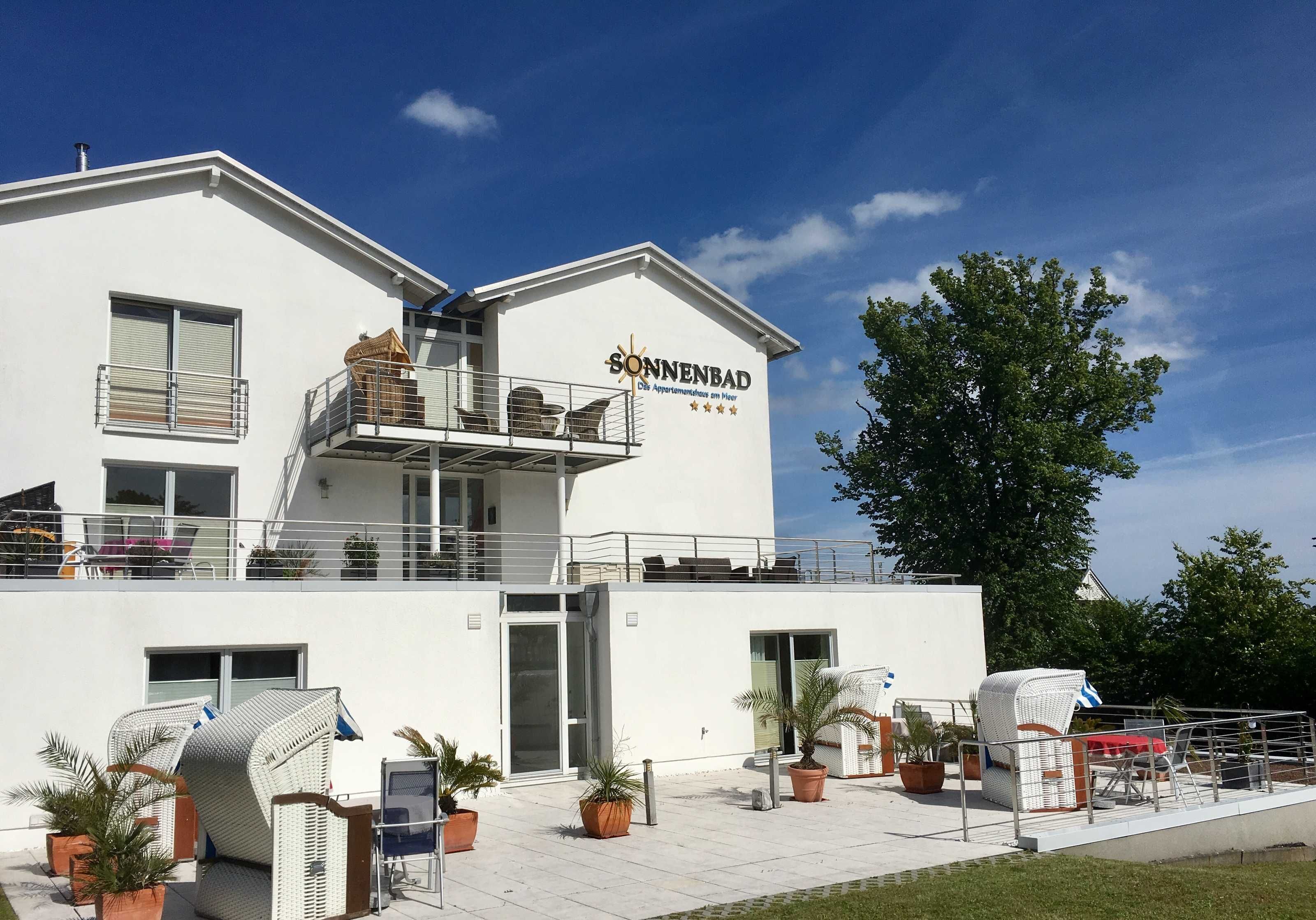 A modern hotel with a white exterior, a balcony with wicker furniture, and potted plants.