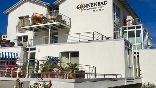 Un edificio moderno con la scritta 'GÖNNENBAD' e una segnaletica che indica si tratta di una residenza con appartamenti. Sono presenti balconi con ringhiere, piante in vaso e mobili da esterno.
