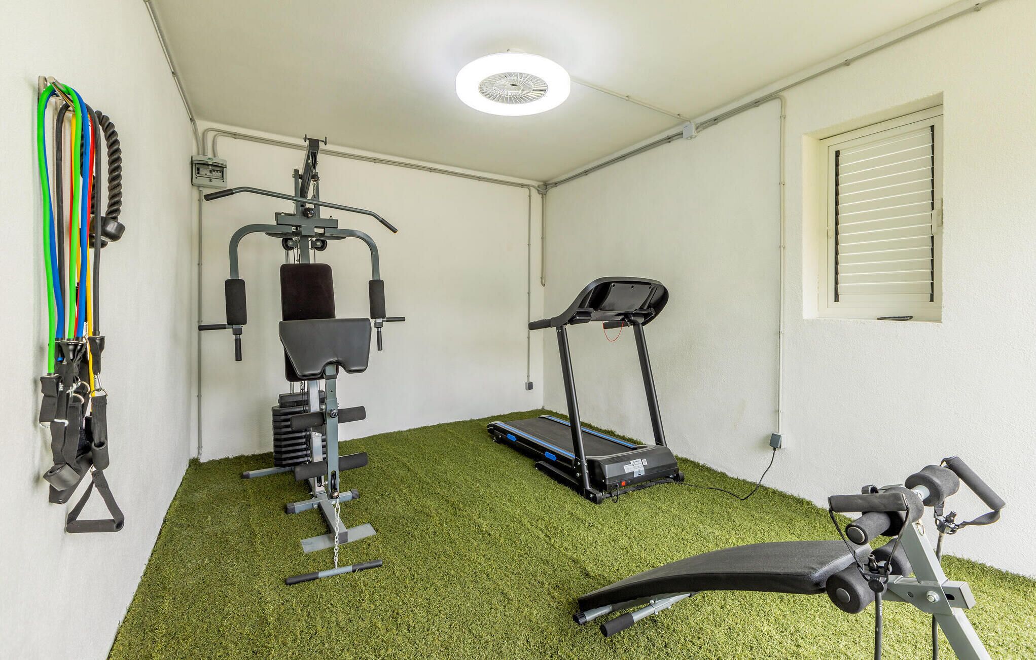 A small, well-lit room with a treadmill, a weight machine, and a bench.