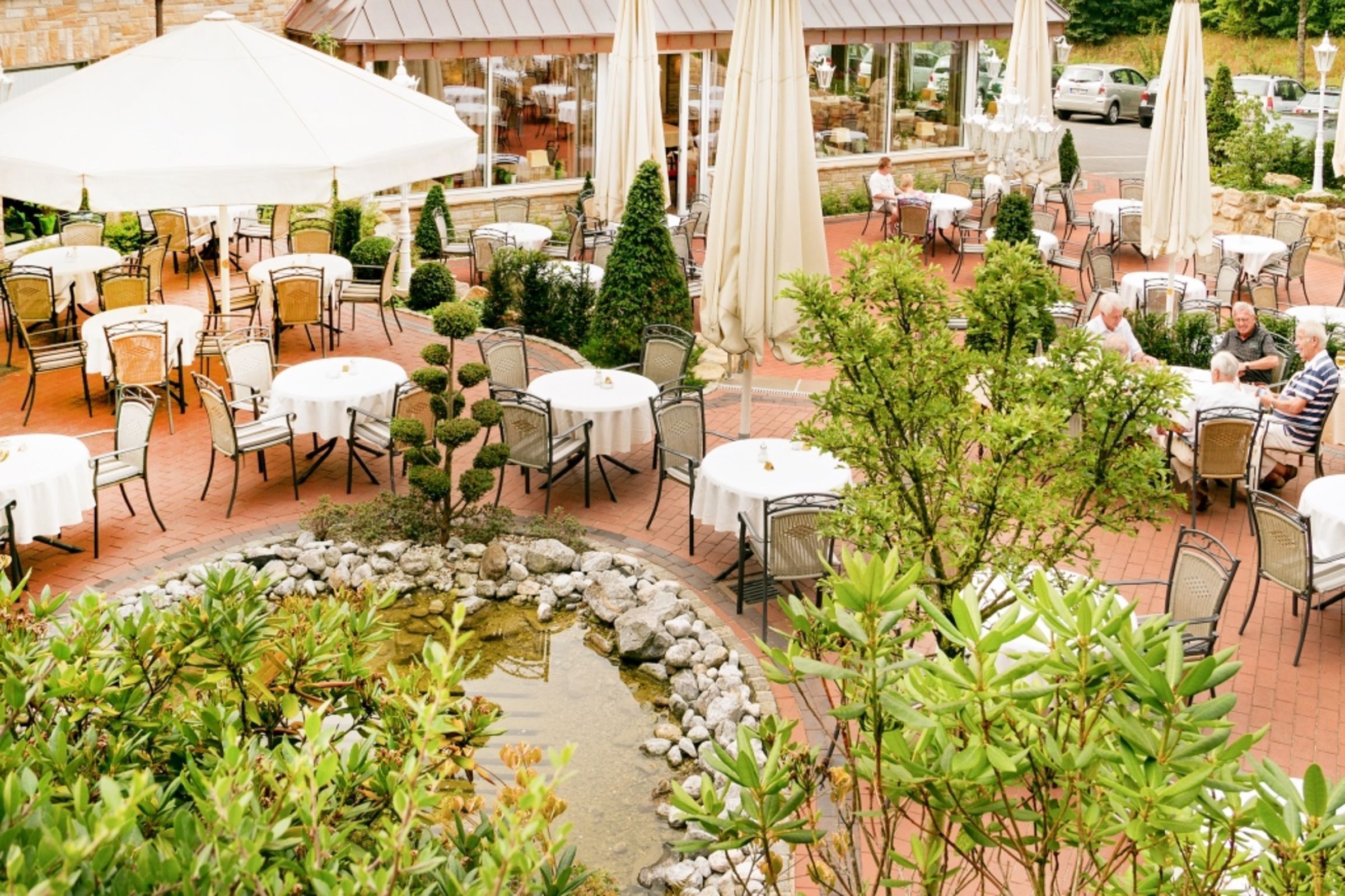 An outdoor patio area with round tables and chairs, surrounded by greenery and a small pond.