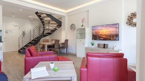 A modern living room with a staircase, a dining area, and a TV displaying a sunset scene.