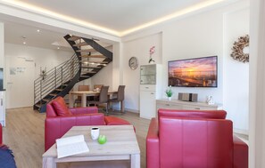 A modern living room with a staircase, a dining area, and a TV displaying a sunset scene. - Lovely apartment in Binz with WiFi (Prora/Rügen)