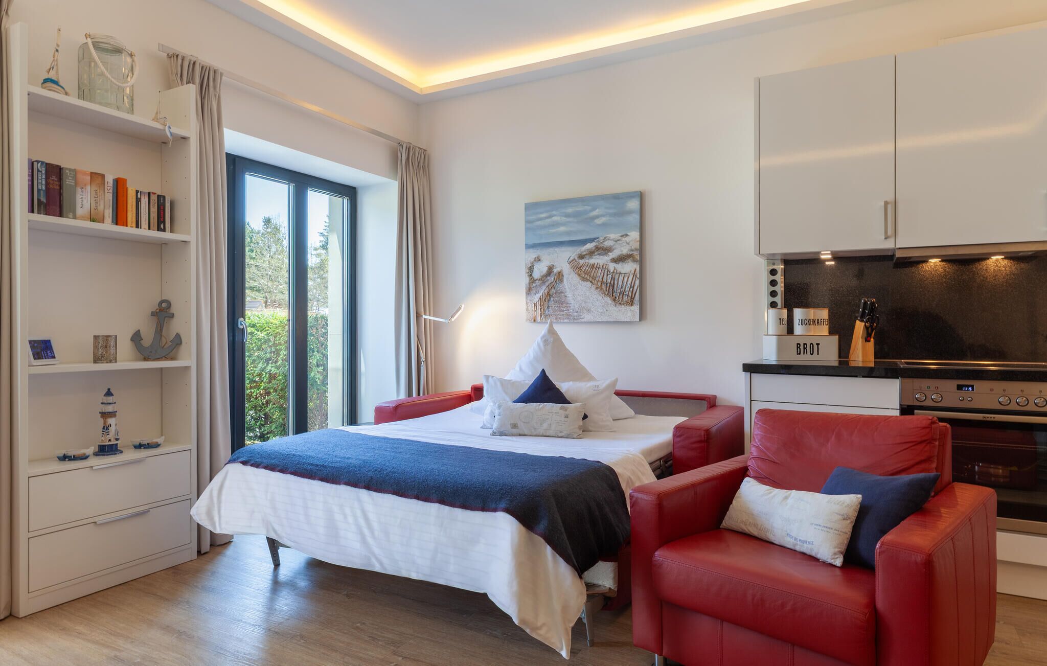 A modern hotel room with a bed, a red armchair, a bookshelf, a painting, and a kitchenette.