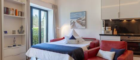 A modern hotel room with a bed, a red armchair, a bookshelf, a painting, and a kitchenette.
