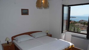 Ein Schlafzimmer mit einem hölzernen Bett, zwei Nachttischlampen, einem kleinen gerahmten Bild und Blick auf das Meer durch ein offenes Fenster.