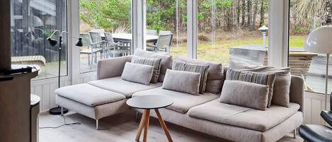 En moderne stue med en hjørnesofa, et rundt sofabord og store vinduer med udsigt til det fri.