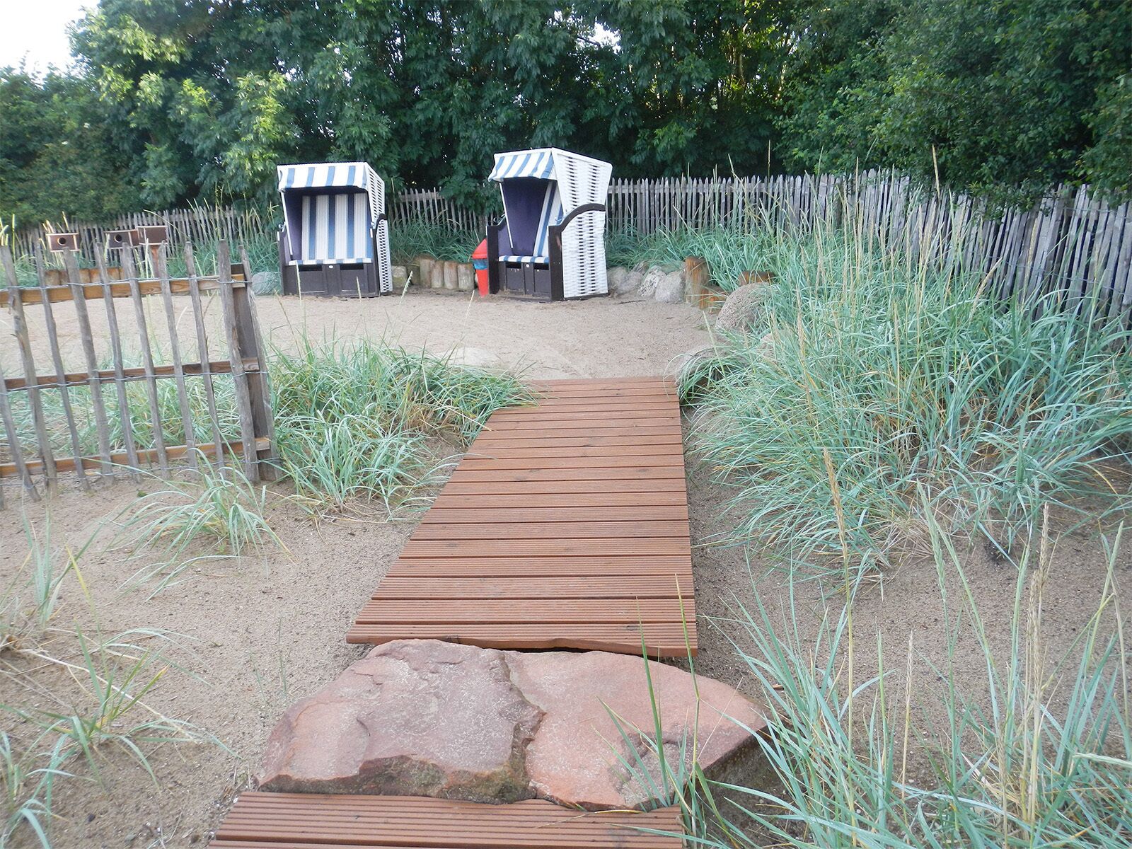 A wooden pathway leading to two striped beach huts with a wooden fence and tall grasses in the background.