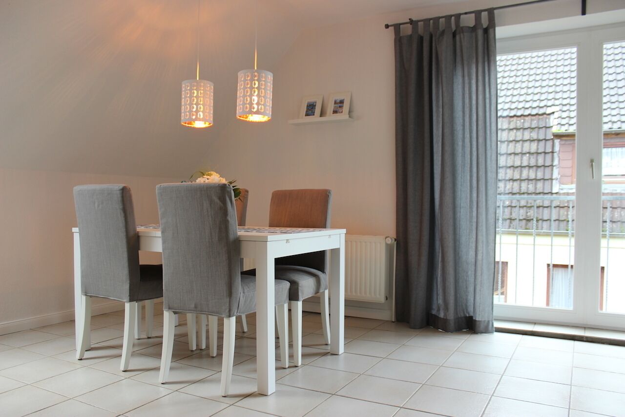 A dining area with a white table and four grey chairs, two pendant lights, a radiator, and a window with grey curtains.