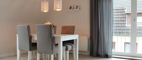 A dining area with a white table and four grey chairs, two pendant lights, a radiator, and a window with grey curtains.