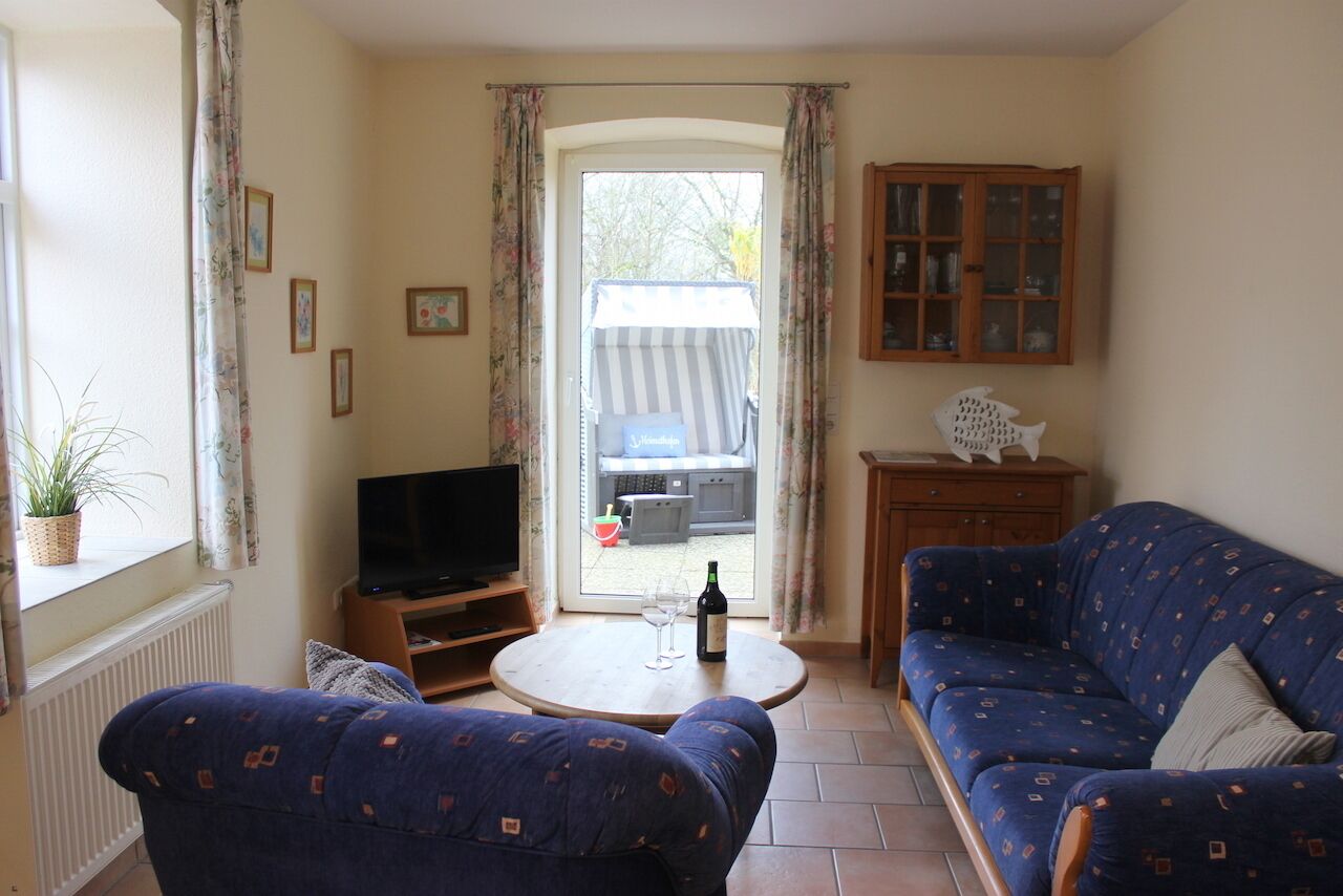 Un salon comprenant un canapé, un téléviseur, une petite table avec une bouteille et un verre, et une vue sur un espace extérieur, visible à travers une porte vitrée.