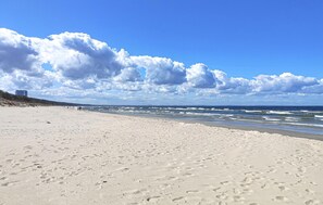 Uma extensa praia de areia com pegadas, céu claro e edifícios ao longe.