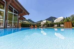 Una piscina con agua azul cristalina, rodeada de sillas de descanso y una terraza de madera, con montañas al fondo.