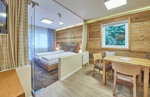 A modern hotel room with a glass partition, a bed, a wooden wall, a window with a view, and a dining area. - Triple in alpine style - Rosentalerhof Hotel & Apartments (Hinterglemm)