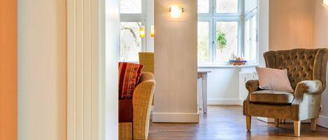 A spacious room with a wooden floor, a large window with a view of greenery, a plush armchair, and a wicker sofa.