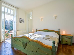 Ein Schlafzimmer mit einem hölzernen Bett, einem Nachttisch, einer Sitzgelegenheit am Balkon und Blick auf Balkonmöbel.
