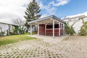 A covered outdoor area with a red wall and white columns, surrounded by a paved patio and a grassy lawn.