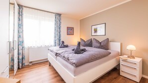 Een slaapkamer met een bed, nachtkastje met lamp, raam met gordijnen en een ingelijst schilderij aan de muur.