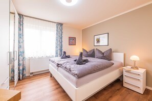 Ein Schlafzimmer mit Bett, Nachttisch mit Lampe, einem Fenster mit Vorhängen und einem gerahmten Bild an der Wand.