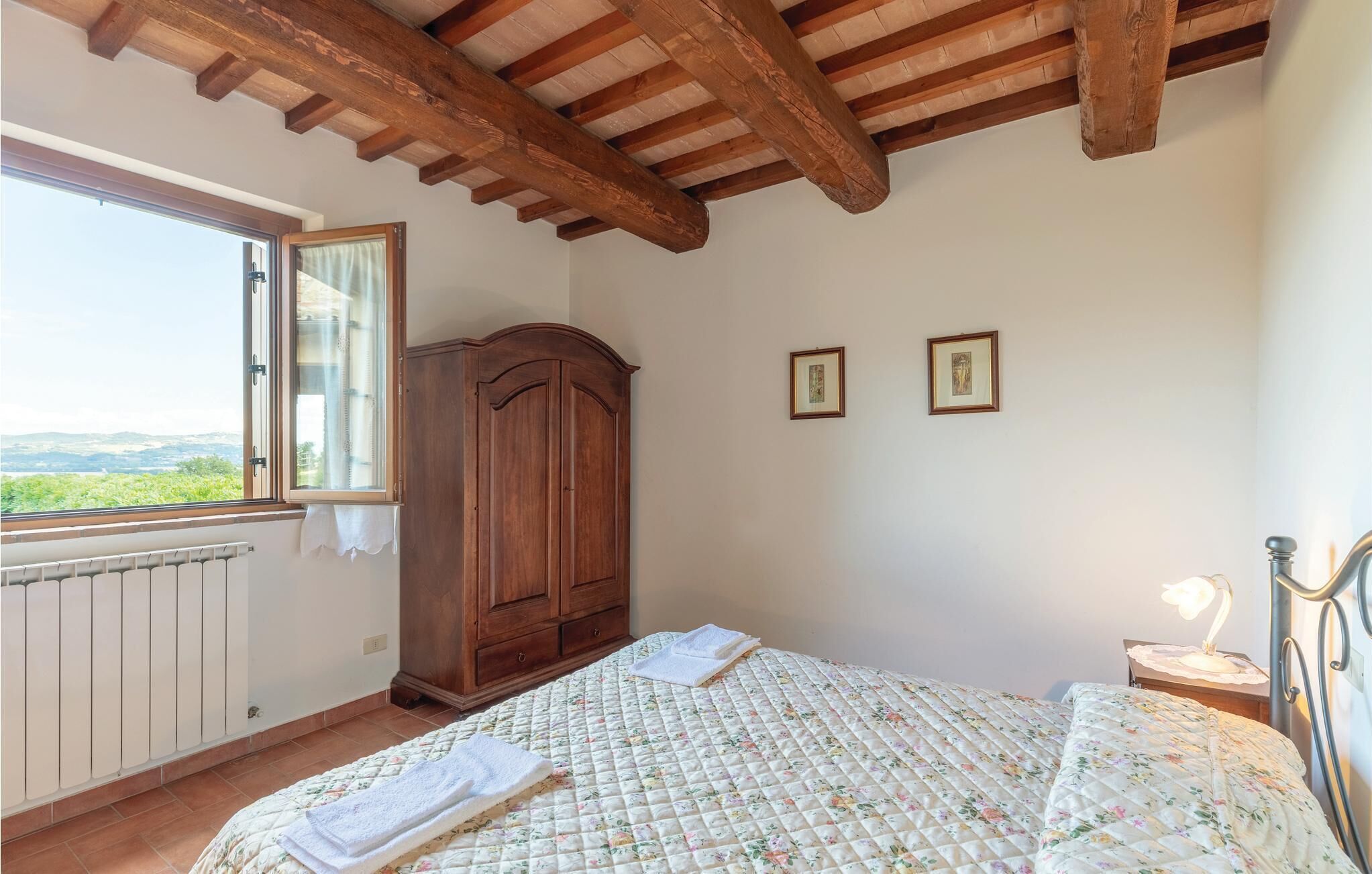 Una camera da letto con soffitto in legno, un'ampia finestra, un armadio, un letto con copripiumino floreale e due quadri appesi al muro.
