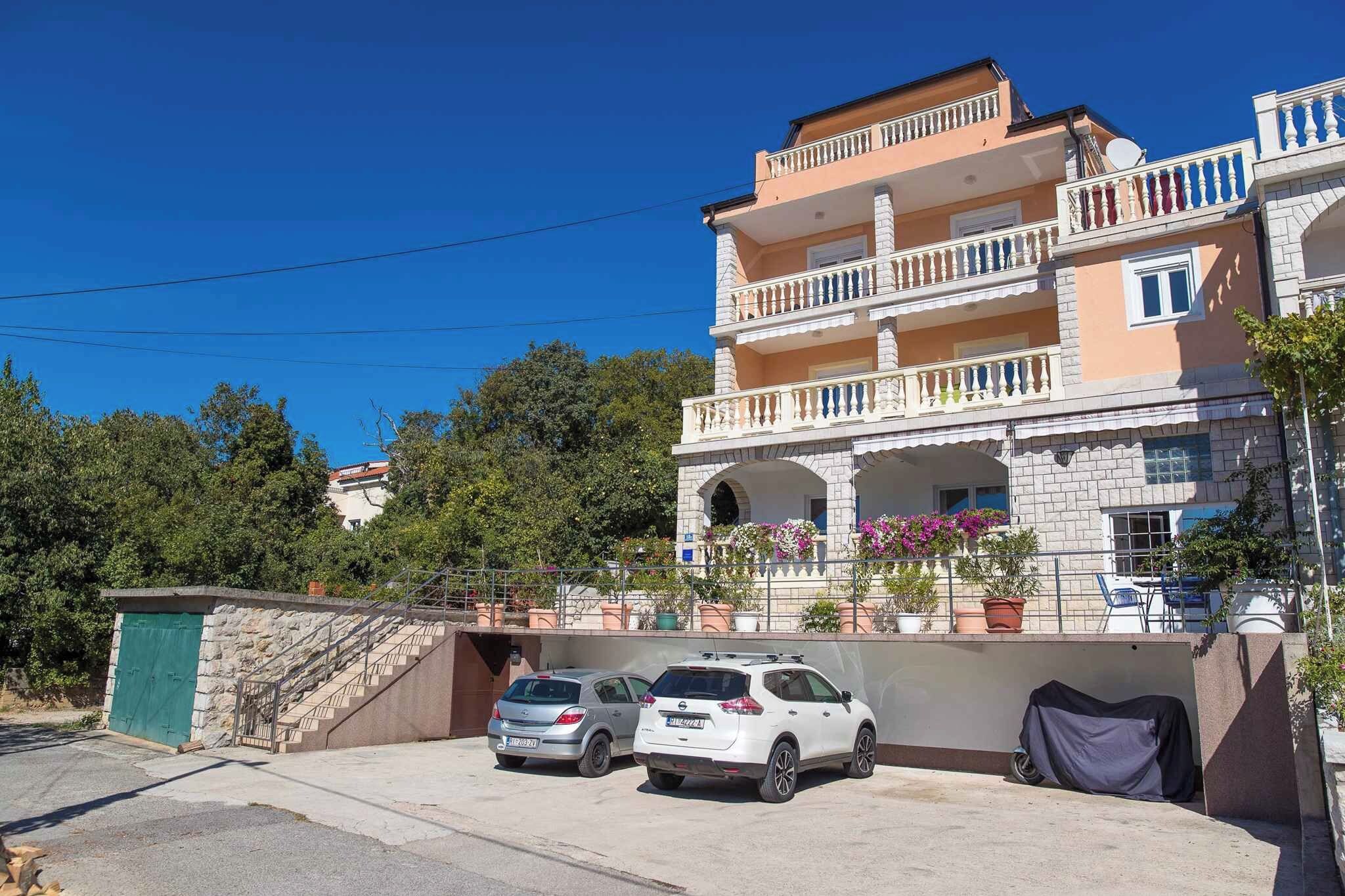 Un edificio residencial de varios pisos con balcones, un estacionamiento con dos coches y plantas en macetas.
