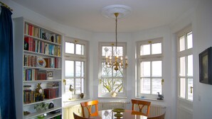 Een kamer met een groot raam, een kroonluchter, een boekenplank en een eettafel met stoelen.