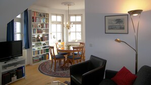 Een woonkamer met een televisie, boekenkasten, een eethoek met stoelen en een vloerlamp.
