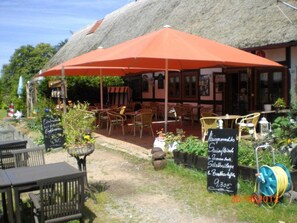 Un espace extérieur aménagé avec des tables et des chaises sous un grand parasol rouge, un tableau noir servant de menu, et un bâtiment à toit de chaume en arrière-plan.