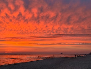 Een strand met een pier die zich uitstrekt in het water, mensen wandelen, en een zonsondergang met oranje wolken.