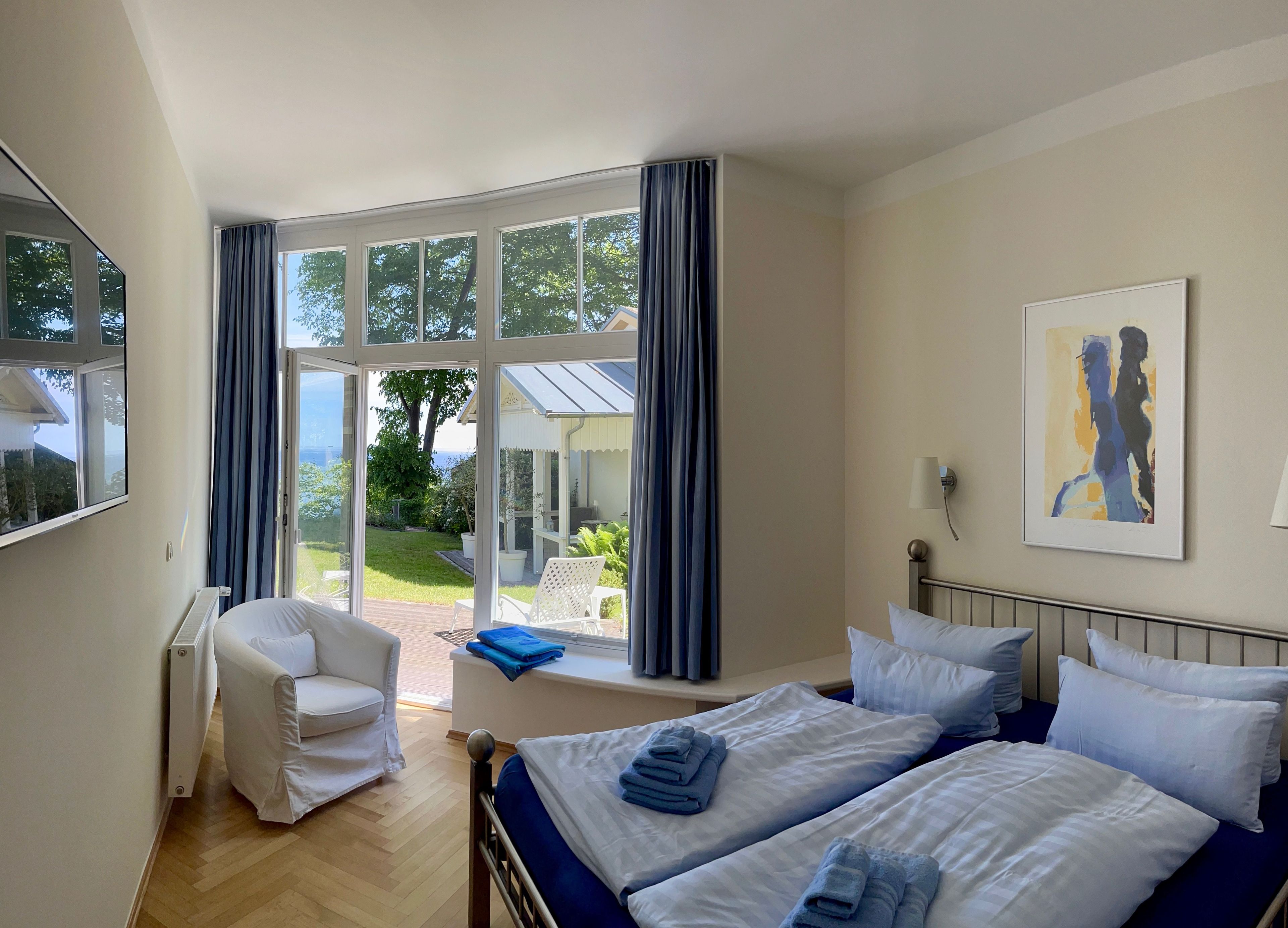 Ein Hotelzimmer mit großem Fenster, einem Bett mit blauer Bettwäsche, einem weißen Stuhl und einem Bild an der Wand.