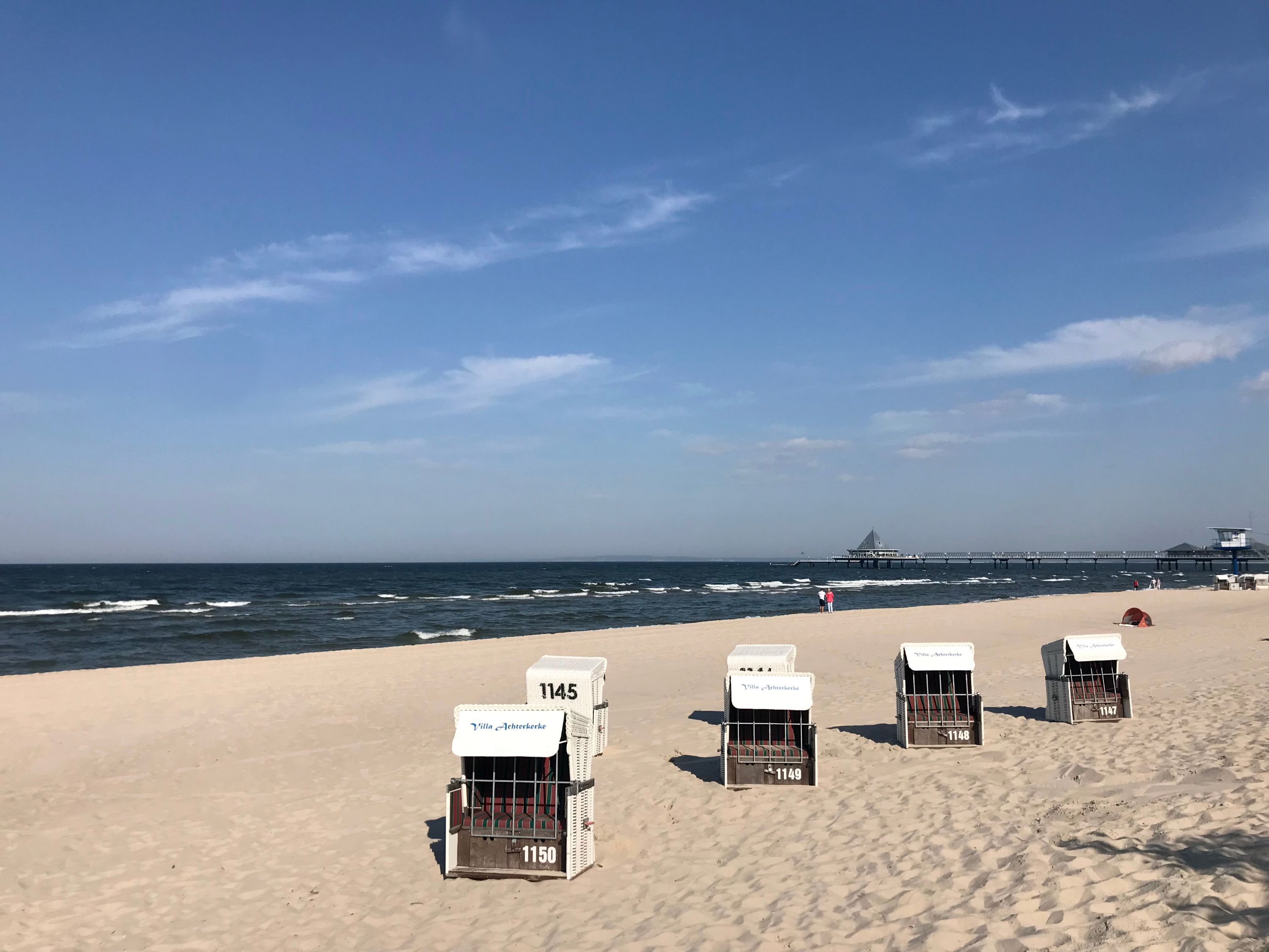 Strandstühle mit Nummern am Strand, ein Steg im Hintergrund und ein klarer Himmel.