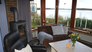 A cozy living room with a fireplace, a sofa, a table with a vase of flowers, and a view of a lake. - RED Holiday home at the Bistensee - Holiday home Frick (Alt Duvenstedt)