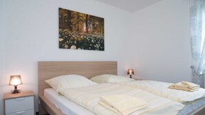 A double bed with a headboard, two bedside tables with lamps, a wall-mounted painting of a forest, and a window with a curtain. - Reriker Straße 06b Whg. RER6b-2 .. - Reriker Str. 6b Whg. RER6b-02 (Kühlungsborn)