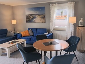 A living room with a blue sofa, a coffee table, a round wooden table with chairs, a painting on the wall, and a window with curtains. - Reriker Straße 06b Whg. RER6b-2 .. - Reriker Str. 6b Whg. RER6b-02 (Kühlungsborn)