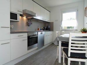 A modern kitchen with white cabinets, a built-in oven, a microwave, and a coffee maker. There is a dining table with chairs and a window providing natural light. - Reriker Straße 06b Whg. RER6b-2 .. - Reriker Str. 6b Whg. RER6b-02 (Kühlungsborn)