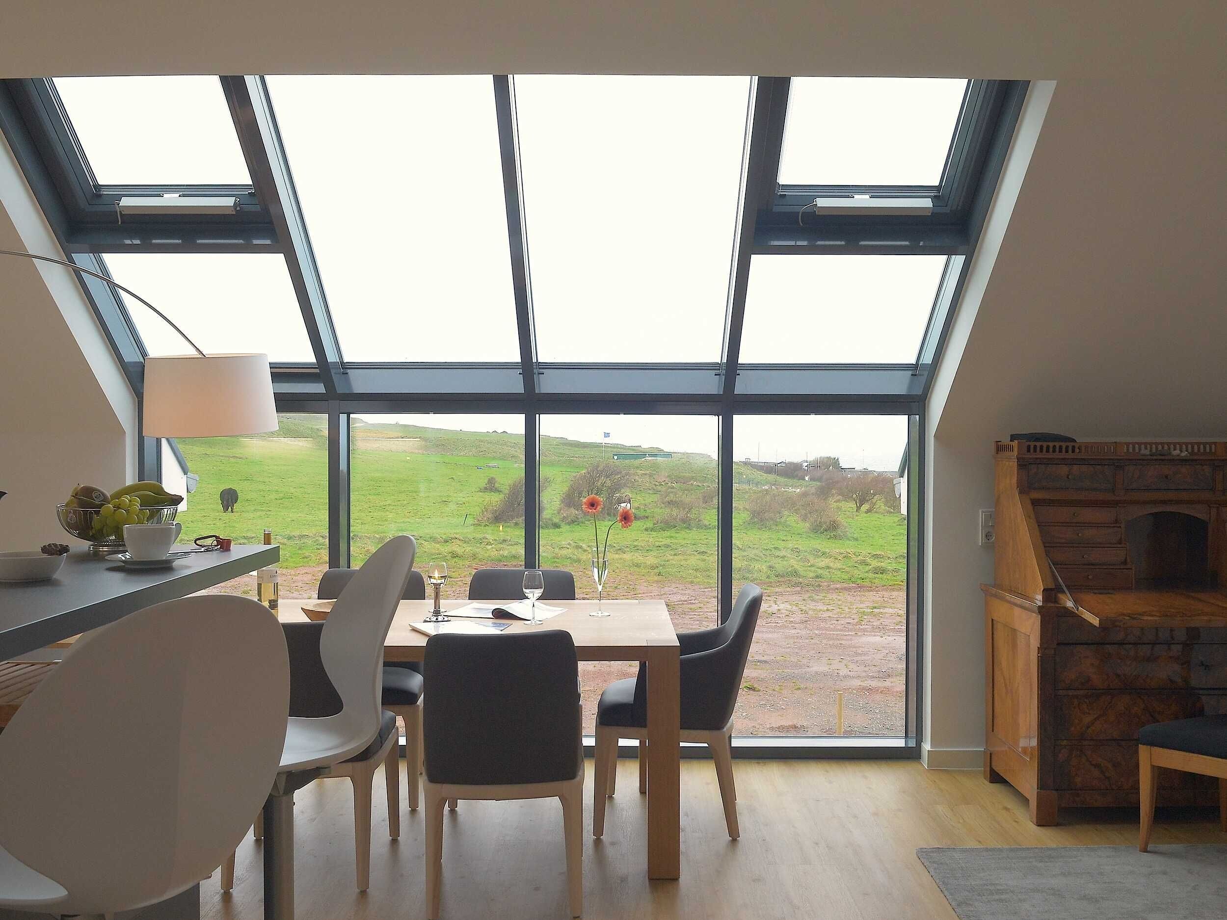 A dining area with a table and chairs, a lamp, and a view of a green landscape through large windows.