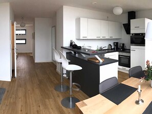 A modern kitchen with white cabinets, a black countertop, and stainless steel appliances. There are two white bar stools and a dining table with a candle.