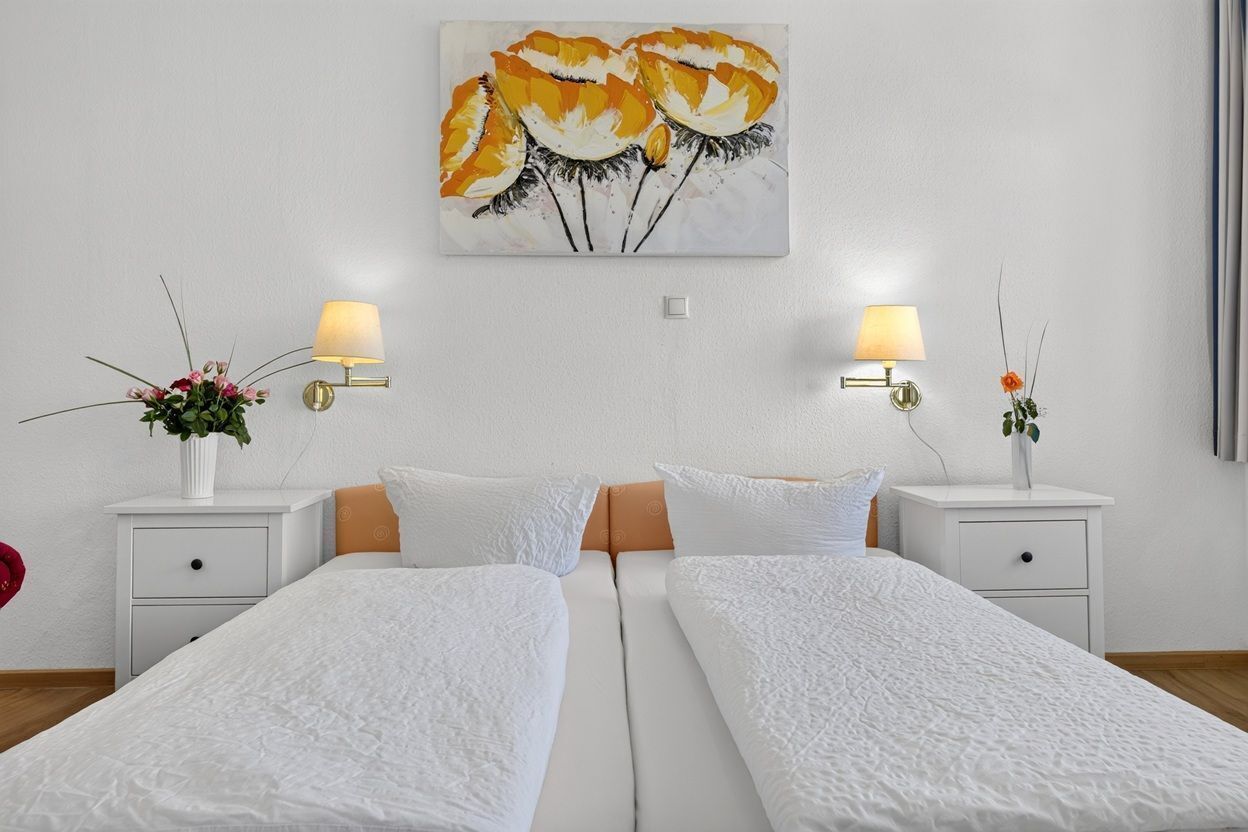 A hotel room with two single beds, a white wall, a painting above the beds, and two bedside tables with lamps and flower arrangements.