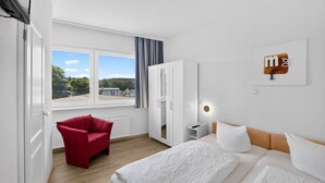 Habitación de hotel con cama, una silla roja, armario, mesita de noche y ventana con vistas a casas.