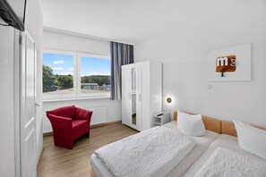 Una camera d'albergo con un letto, una poltrona rossa, un armadio, un comodino e una finestra vista case.