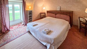 A bedroom with a large bed, a wooden headboard, a nightstand, a lamp, a small table, and a view of greenery through the window.