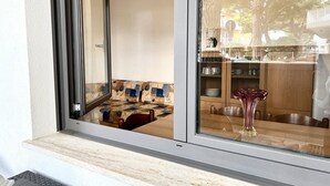 Ein Zimmer mit einem großen Fenster, einer hölzernen Bank und einem durch das Fenster einsehbaren Küchenbereich.