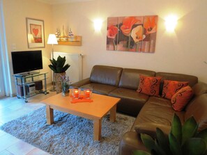 A living room with a brown leather sofa, a glass coffee table, a flat-screen TV on a stand, a potted plant, and a wall-mounted painting.