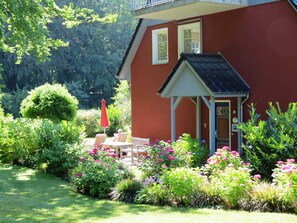 A red building with a small porch and a garden with flowers and shrubs.