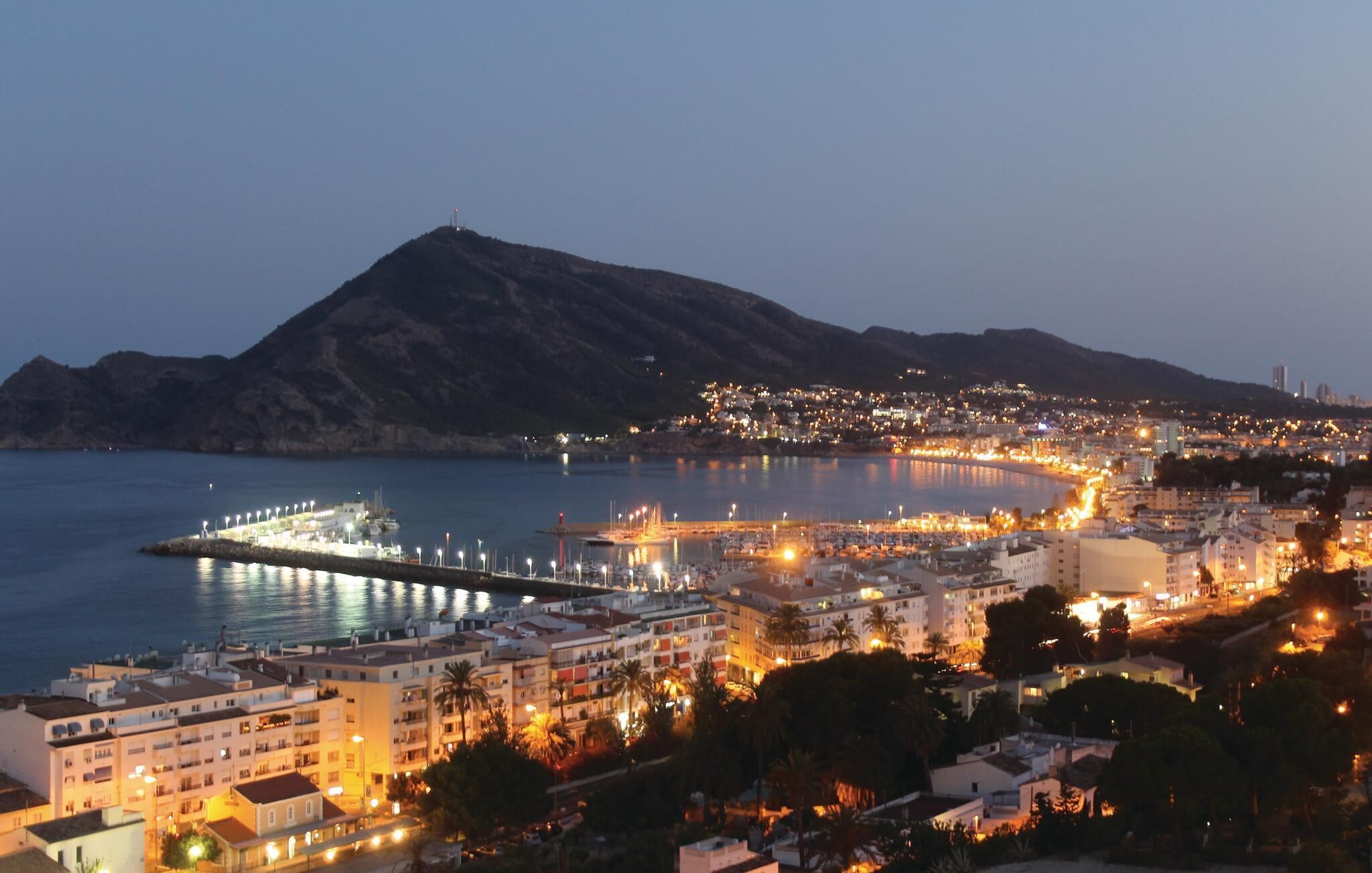 A coastal cityscape at dusk with a prominent pier extending into the sea, surrounded by residential buildings and a mountain in the background.