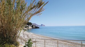 Een strand met helderblauw water, een kiezelstrand en rotsformaties in de verte.