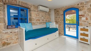 Una stanza con parete in pietra, una finestra con cornice blu, un letto con biancheria azzurra e un comò bianco.