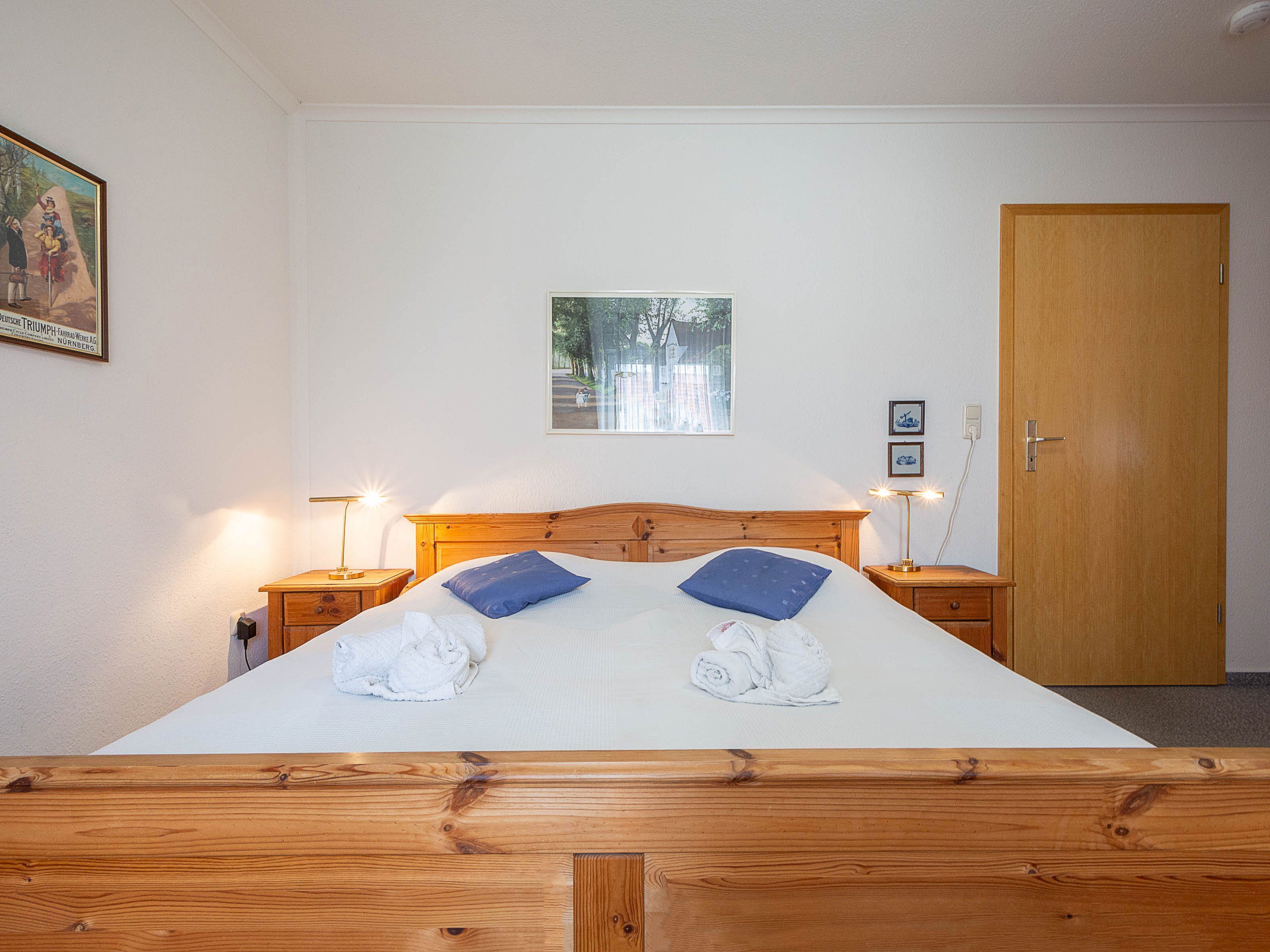 A neatly made bed with two pillows and two folded towels, a wooden headboard, two bedside tables with lamps, a framed picture on the wall, and a door to the right.