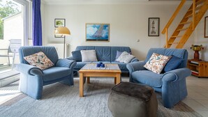 A living room with two blue sofas, a wooden coffee table, and a grey ottoman. There is a staircase to the right and a window with curtains on the left. - Reriker Straße RER20-02. - Reriker Straße 20 Whg. 02 (Kühlungsborn)
